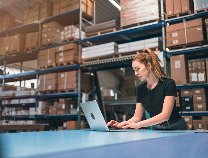A woman using a laptop in a warehouse, focused on managing an operating lease for asset finance